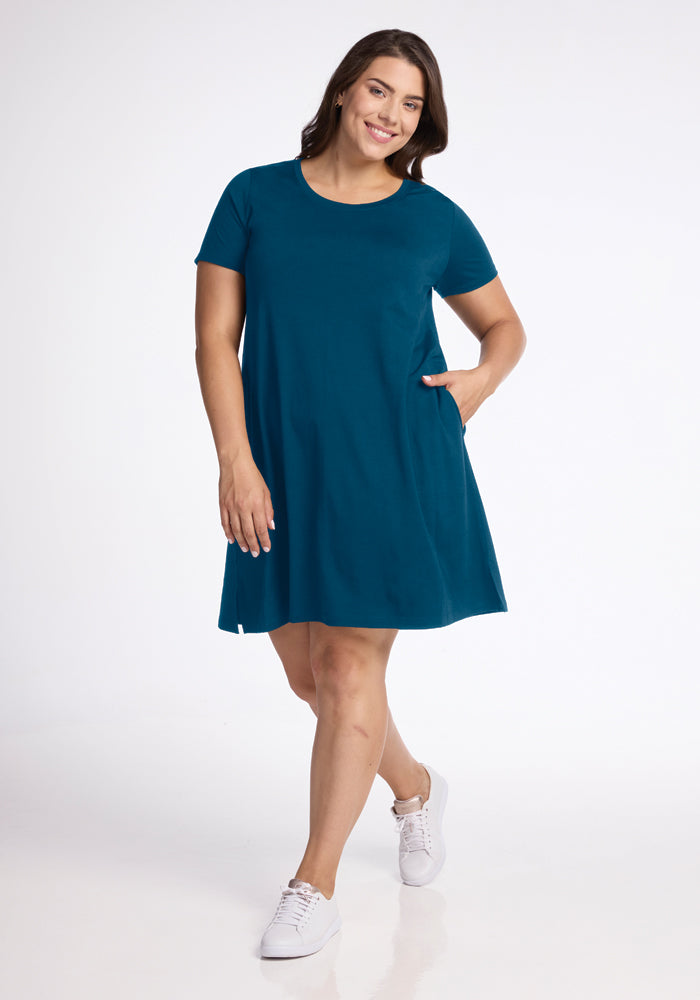 A person wearing the Woolx Georgie Dress in Real Teal, crafted from lightweight fabric, smiles as they walk. They're paired with white sneakers and have one hand casually placed in their pocket against a plain white background, emphasizing the relaxed look.