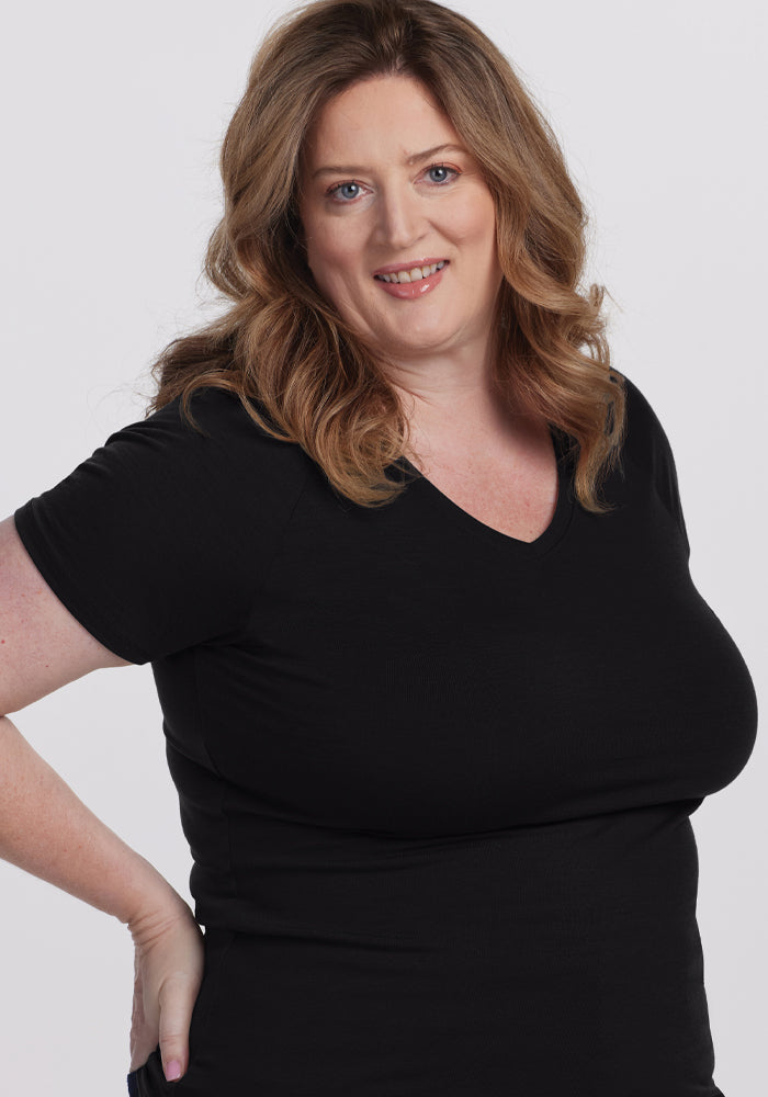 A person with long, wavy brown hair smiles at the camera while wearing the Woolx Mia V Neck - Black. The plain white background highlights the garment's superior quality.