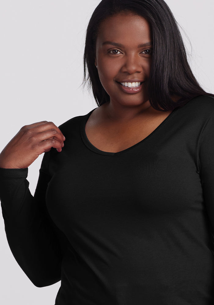 A smiling woman with long dark hair, wearing the Woolx Layla V Neck Top in black, poses effortlessly against a light gray background. Her left hand near her shoulder showcases the top's stylish and functional design in one graceful gesture.