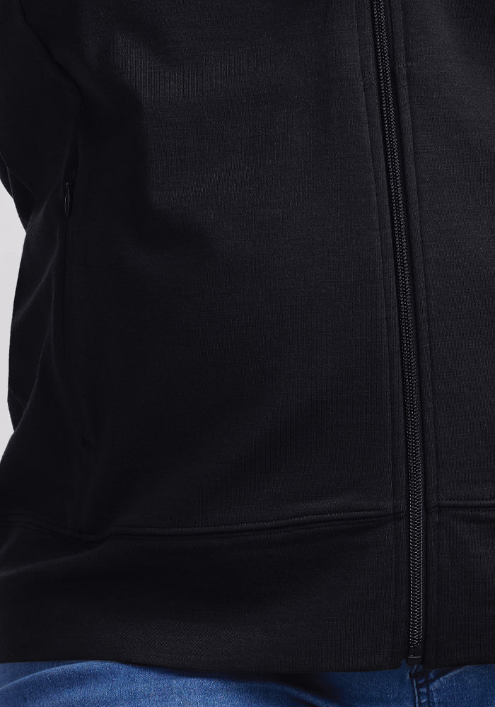 The image shows a close-up of someone in blue jeans wearing the Woolx Ryann Hoodie in black, highlighting its sleek design and secure zippered pockets.