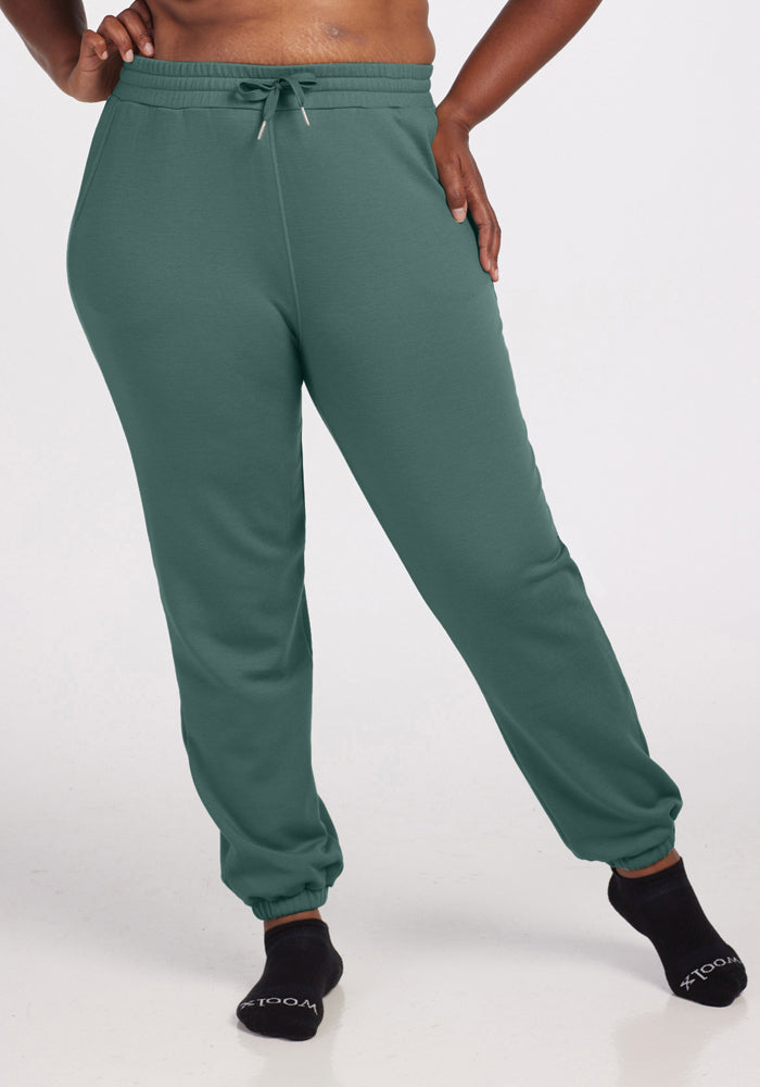 A person wearing Woolx's Parker Sweatpants in Duck Green and black socks is standing with one leg slightly bent. The outfit looks comfortable and casual, ideal for lounging or exercising. The plain, light-colored background enhances the relaxed atmosphere. 