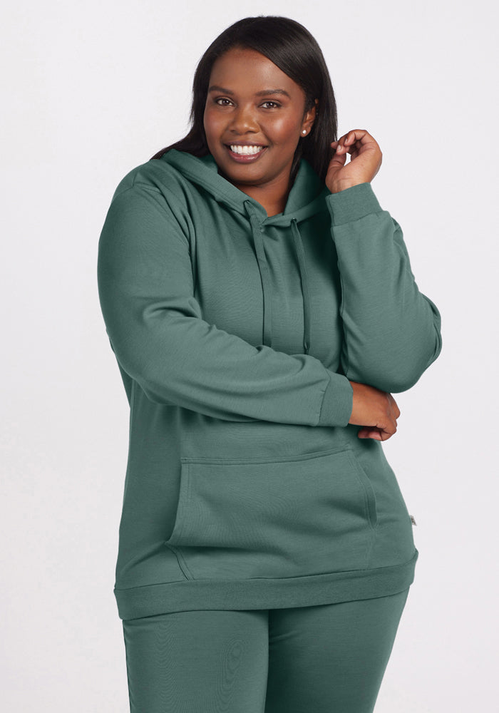 A person with long dark hair, smiling and exuding warmth and style, stands against a plain background wearing a Duck Green Avery Hoodie from Woolx, part of the WoolAire Collection™. Paired with matching sweatpants, the luxurious Merino wool hoodie has their hands tucked in its pockets. 