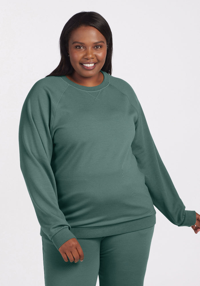 A person smiling while wearing a sustainable Bailey Crewneck Sweatshirt in Duck Green from Woolx, paired with matching pants from the Wool Aire Collection, standing against a plain background. 