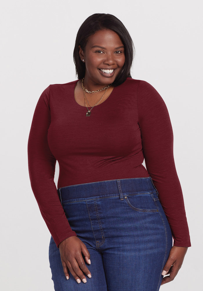 A smiling woman with dark hair stands against a plain background, exuding a chic look in a maroon Sophia Bodysuit by Woolx paired with high-waisted blue jeans. She rests her hand on her hip and accessorizes with a layered necklace. 