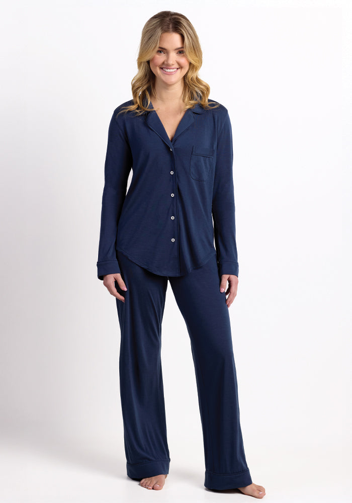 A person stands smiling, wearing the Harper FeatherTouch® Pajama Set - Starry Night by Woolx, a matching navy blue ensemble made of breathable Merino wool featuring a button-up top and long pants. The plain white background highlights the comfort and moisture-wicking qualities of their attire.