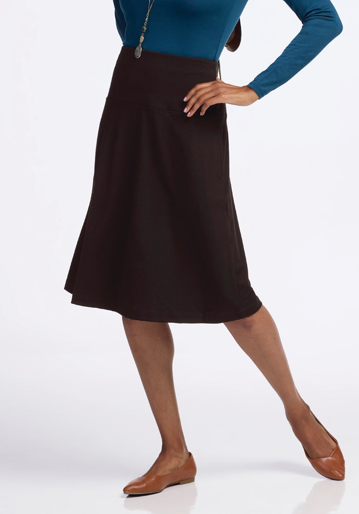 A person dressed in a blue long-sleeve top and the Marilyn Flowy Midi Skirt in French Roast by Woolx stands against a white background. They are wearing brown flats and have one hand on their hip.