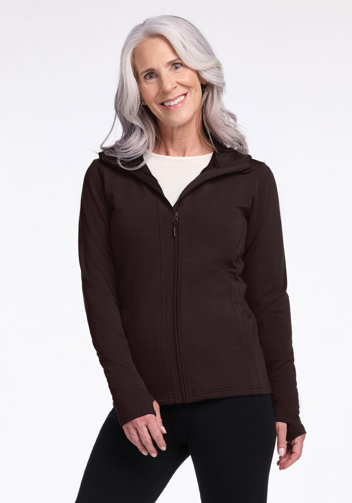 A smiling woman with long white hair is wearing a Woolx Zoey Zip Up Hoodie in French Roast over a white shirt. Perfect for layering, the zip-up hoodie is made from luxurious Australian Merino Wool. She is standing against a plain white background. 