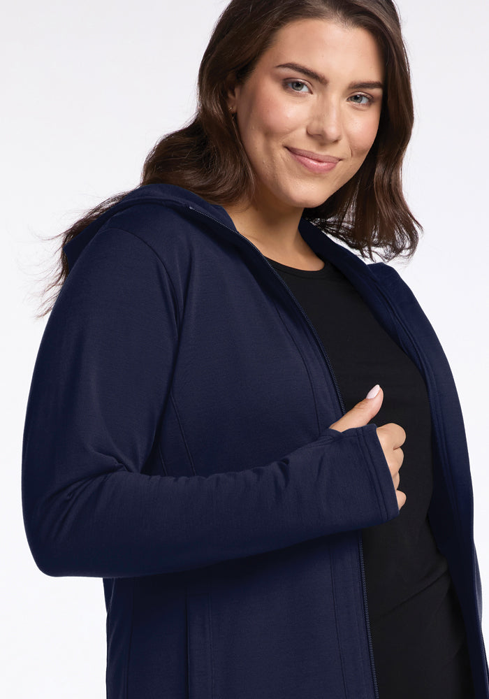 A person with long brown hair is smiling and wearing a Woolx Zoey Zip Up Hoodie in Deep Navy over a black shirt. The background is plain and light-colored. The individual's left hand is partially visible, giving a thumbs-up gesture.