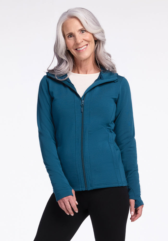 A woman with long, gray hair is smiling at the camera. She is wearing a Zoey Zip Up Hoodie in Real Teal by Woolx and black pants made from Woolx DuraLite fabric. The background is plain white. 