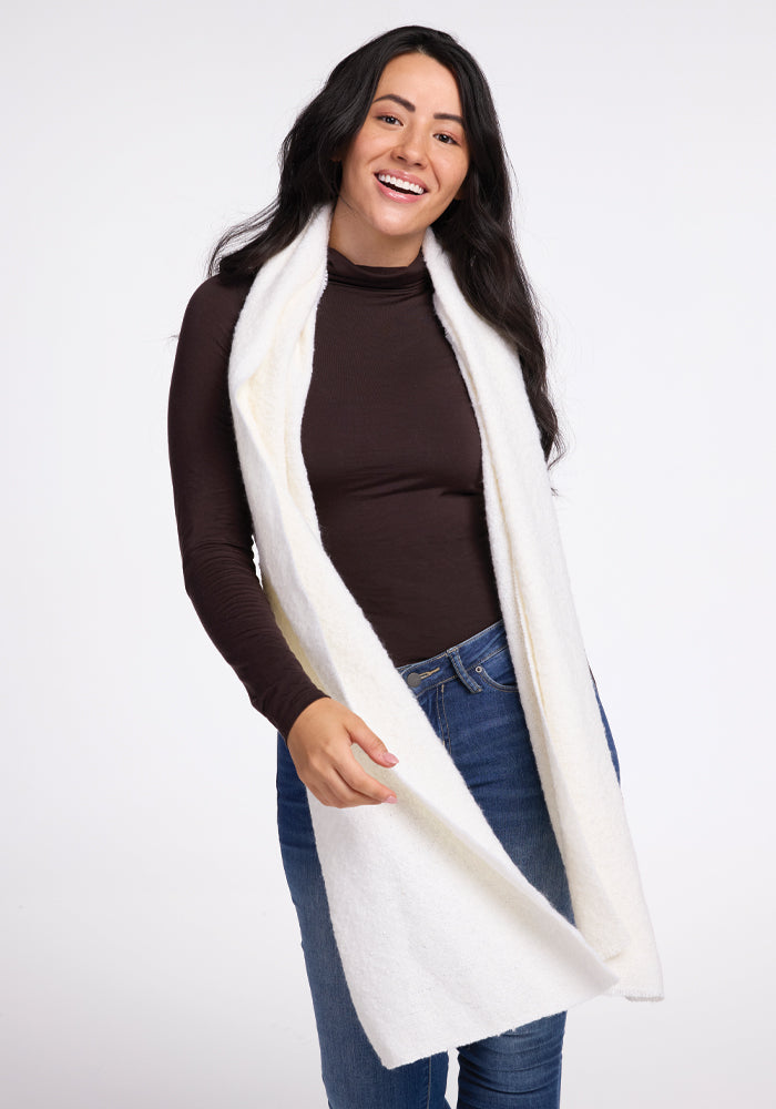 A person with long dark hair is smiling and wearing a black turtleneck, blue jeans, and the Bradie Scarf in cream by Woolx. The background is plain white.