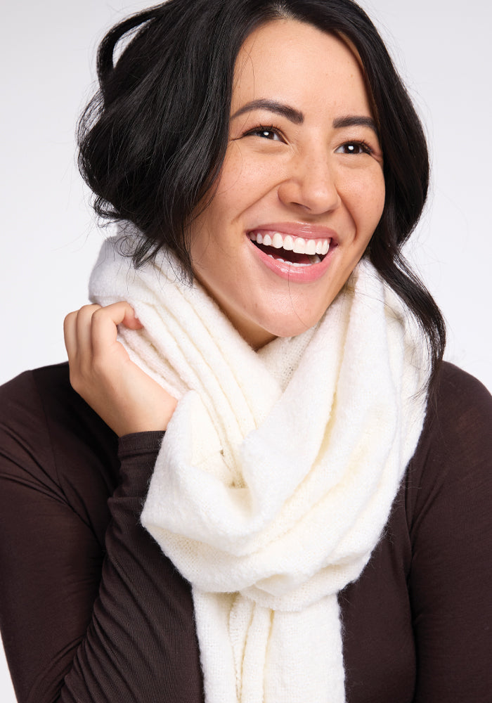 A person with long dark hair, wearing the Bradie Scarf in Cream by Woolx and a brown top, is smiling brightly against a plain background. Their hand is lightly touching the scarf, creating a joyful and cozy atmosphere.